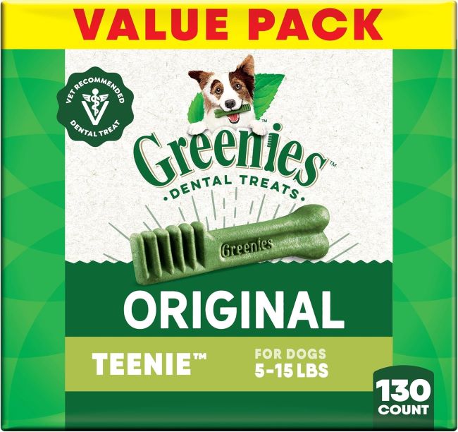 A happy medium‑sized dog joyfully chewing a GREENIES Original Natural Dental Dog Treat, with the bright green packaging (130‑count) visible in the background.